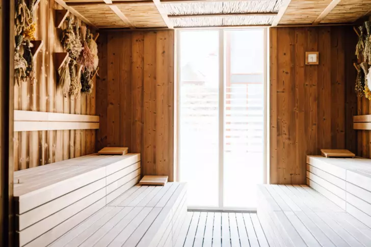 Eine holzverkleidete Sauna mit Blick auf ein teils verdecktes großes Fenster durch das sonniges Tageslicht hereinscheint. An der wand hängen getrocknete Kräuter. 