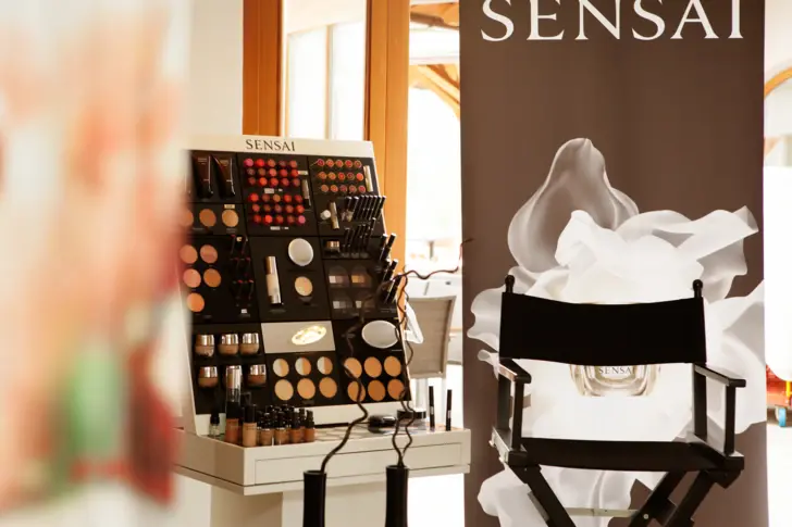 Zur linken Seite des Bildes steht ein Make Up Aussteller von SENSAI mit unterschiedlichsten Produkten. Daneben steht ein schwarzer Stylingstuhl. 