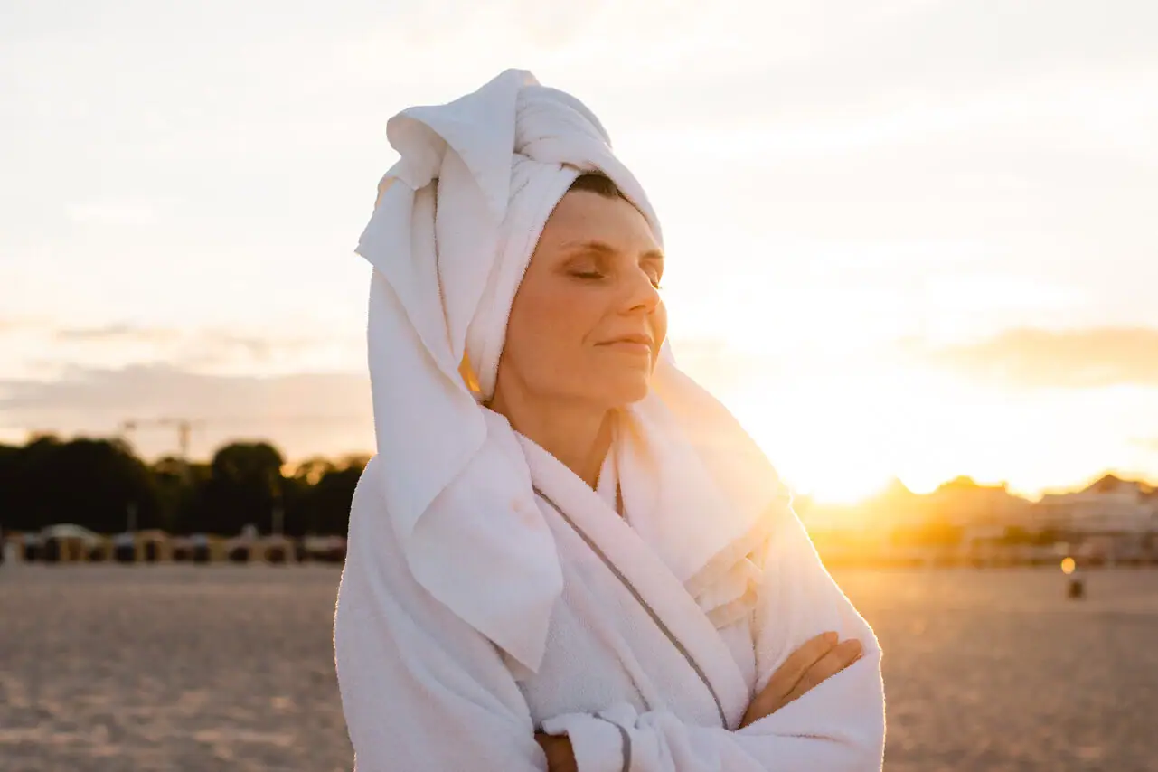 Eine Frau im Bademantel mit einem Handtuch auf dem Kopf steht am Strand.