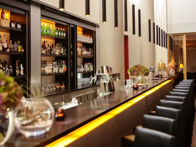 Elegante Bar im A-ROSA Hotel auf Sylt mit einer beeindruckenden Auswahl an Spirituosen auf beleuchteten Regalen, stilvoller Beleuchtung von der Decke und einer modernen Theke mit gelbem Lichtakzent, die eine warme und einladende Atmosphäre schafft. Die Barhocker stehen bereit für Gäste, um einen entspannten Abend mit exquisiten Getränken zu genießen.