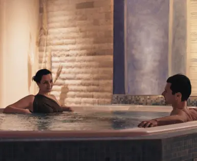  Ein Paar genießt gemeinsam wohltuende Momente im warmen Whirlpool, umgeben von stimmungsvoll beleuchteten Natursteinwänden.