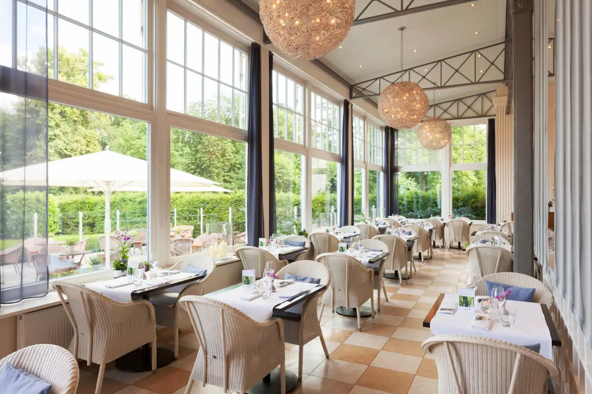 Das Wintergarten-Restaurant im A-ROSA Travemünde mit vielen hellen Korbsesseln und eingedeckten Tischen. 