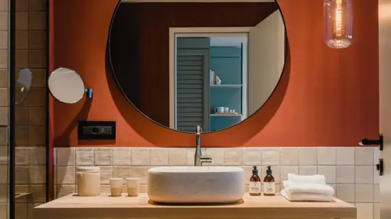 A tastefully furnished bathroom with a terracotta-colored wall. In the center is a washbasin with a large round mirror light above it. A modern hanging pendant light illuminates a white oval washbasin on a light-colored wooden shelf. Next to the washbasin are stacked towels and elegant care products that emphasize the luxurious character of the room. The mirror reflects an open door, offering a glimpse into another room.