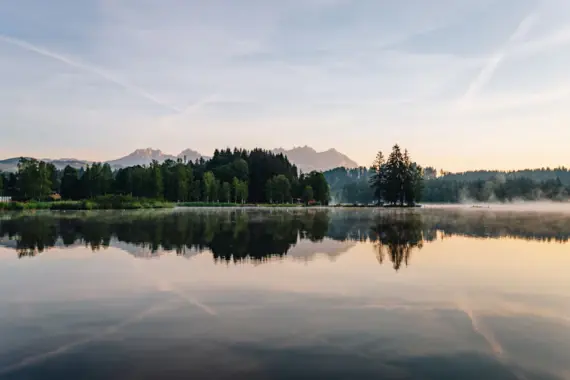 Ein See umgeben von Bäumen mit Bergen im Hintergrund und Spiegelungen auf der Wasseroberfläche.