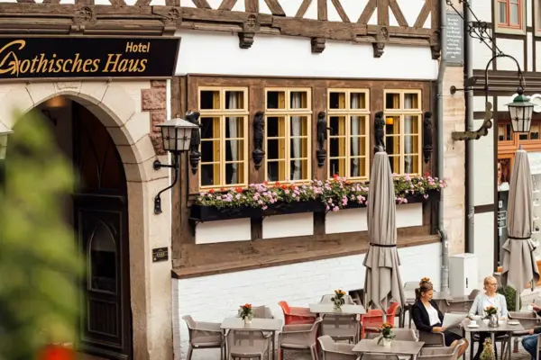 Außenansicht des Hotels Gothisches Haus in Wernigerode mit gemütlicher Terrasse und Fachwerkfassade. Gäste genießen bei Blumen und Sonnenschirmen Kaffee in historischer Altstadtatmosphäre.