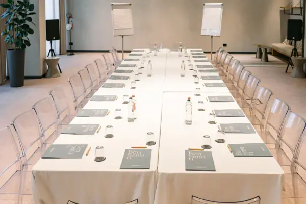 Ein langer Tagungstsch mit weißem Tuch und Gläsern darauf. Außerdem stehen Wasserflaschen auf dem Tisch und es sind Stühle um den Tisch platziert. Im Hintergrund sieht man zwei Flipcharts und Lautsprecher.