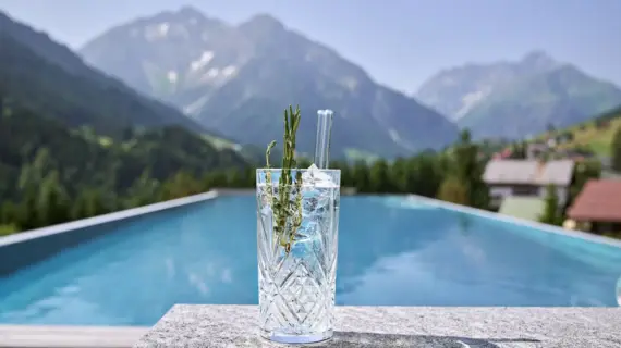 Auf einem Tisch am Pool steht ein Gin Tonic. Im Hintergrund erstreckt sich die Bergwelt. 