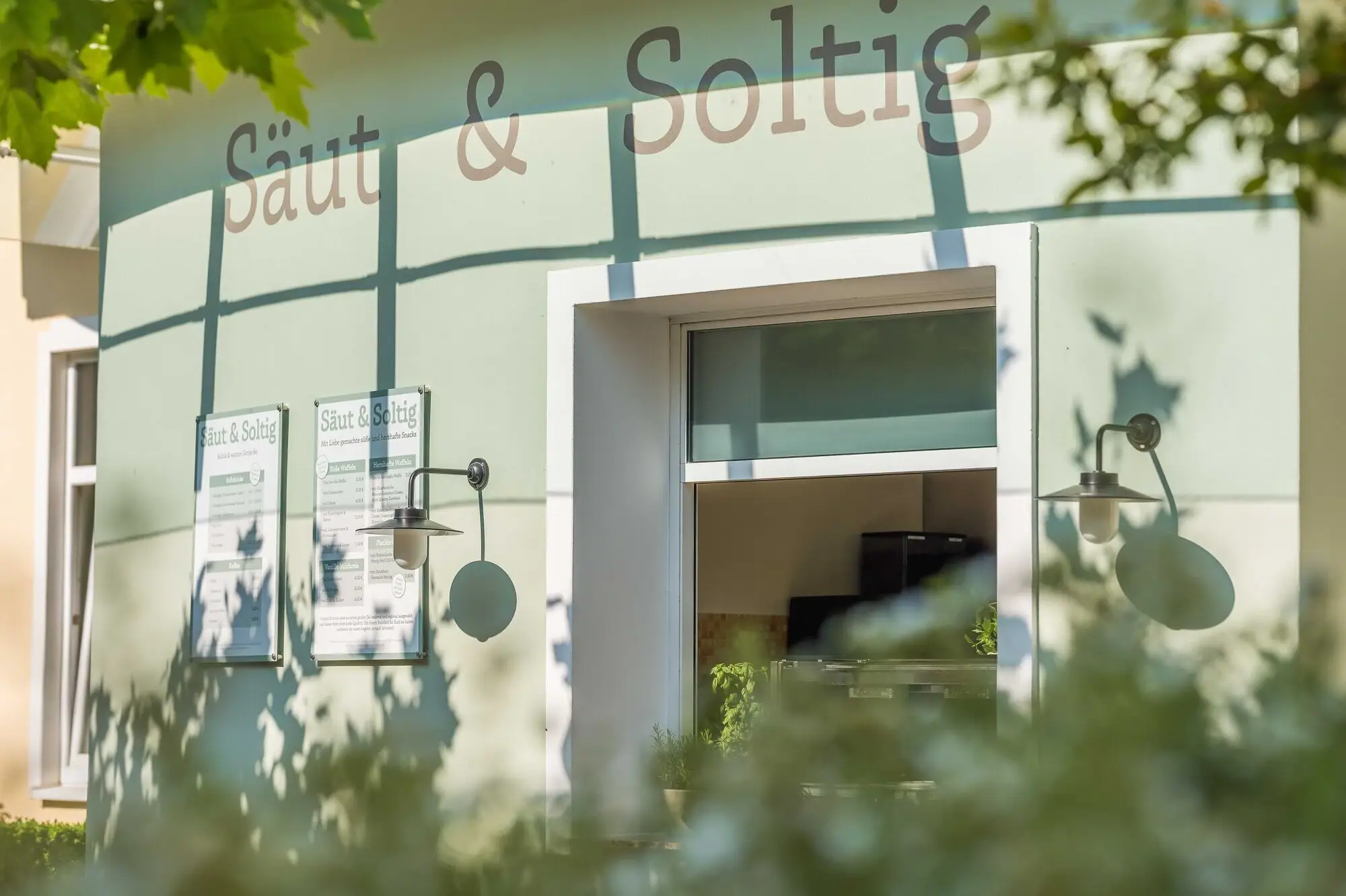 Außenansicht des Bistros „Säut & Soltig“ mit Sonnenschein und grünen Blättern