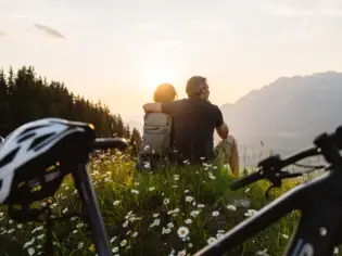 Ein Paar sitzt auf einem Berg und schaut in den Sonnenuntergang während im Vordergrund ein Fahrrad mit einem Fahrradhelm auf dem Sattel steht. 