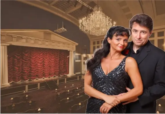 Ein elegant gekleidetes Paar posiert in einem festlichen Theater mit rotem Vorhang, Kronleuchtern und gedämpfter Beleuchtung.