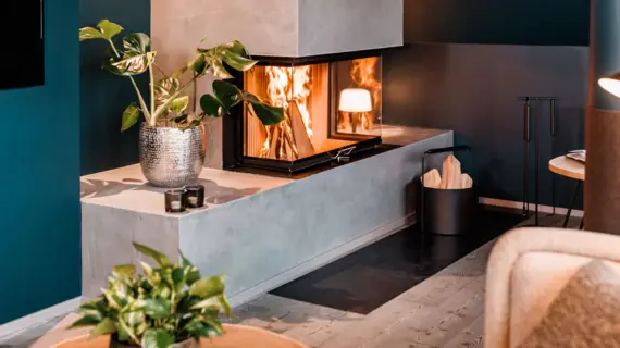 A fire burns in a modern fireplace and in front of it is a container of wood and a plant. 