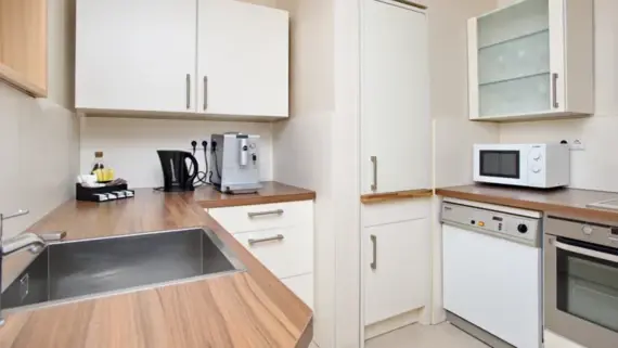 Eine Küche in U-Form mit einer hellbraunen Arbeitsplatte und weißen Schränken. Rechts ist ein Waschbecken, eine Teestation sowie eine Kaffeemaschine zu sehen und links der Kühlschrank, eine Mikrowelle, eine Spülmaschine und ein Backofen.