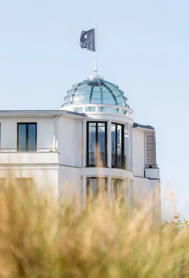A flag with the inscription "CERES" flies in the wind on a glass dome on a white building. The grasses of the dunes can be seen in the foreground.