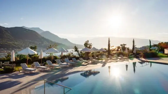 Pool mit Sonnenschirmen und Liegestühlen umgeben von Bäumen und Bergen mit Blick auf den Gardasee.