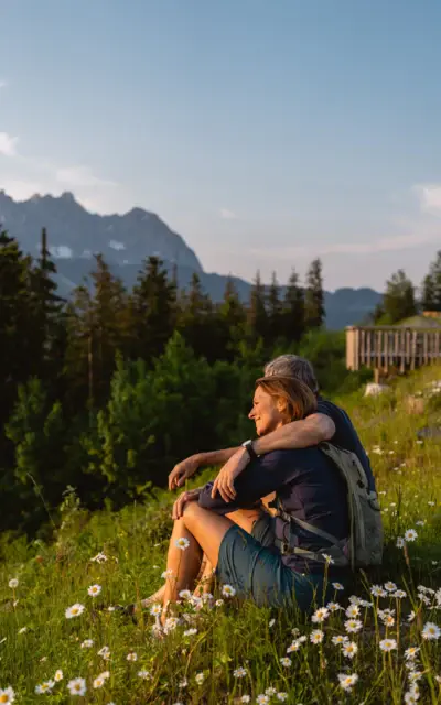 Ein Mann und eine Frau sitzen auf einem Hügel, umgeben von Bäumen und Bergen im Hintergrund.