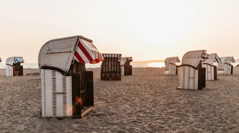 Strandkörbe mit blau und rot weißem Sonnenschutz stehen entlang eines Strandes verteilt. Das Meer schimmert von der aufgehenden Sonne, welche die Szene in warmes Licht taucht.