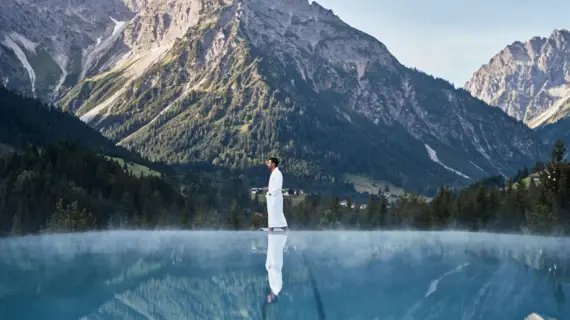 Ein Mann in Bademantel spaziert einen Beckenrand am Pool entlang mit Blick auf die im Hintergrund liegende Bergwelt. 
