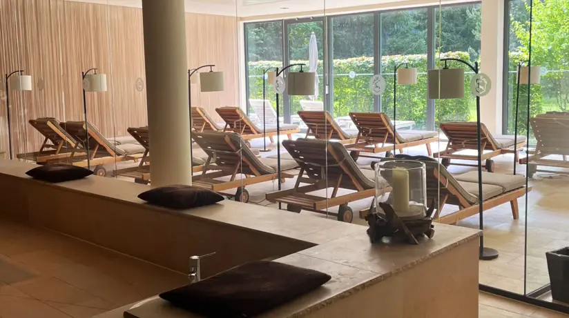  Ein eleganter und ruhiger Ayurveda-Behandlungsraum mit sanfter Beleuchtung. In der Mitte des Raumes befindet sich eine Massageliege, liebevoll vorbereitet mit einem Handtuch und Rosenblättern. Im Hintergrund steht eine moderne, freistehende Badewanne neben einem Fenster, das mit warmtonigen Vorhängen versehen ist. Künstlerische Objekte und Kerzenständer verleihen dem Raum eine beruhigende und luxuriöse Atmosphäre.