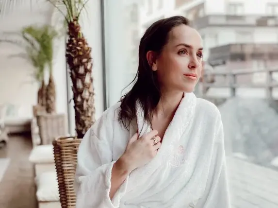 A woman in a white bathrobe sitting by a window.