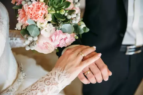Nahaufnahme der Hände eines frisch verheirateten Paares, die Eheringe tragen und sich halten, mit einem wunderschönen Hochzeitsstrauß aus rosa Blumen und grünen Blättern im Hintergrund. Die Braut trägt ein Kleid mit Spitzenärmeln, der Bräutigam einen schwarzen Anzug mit weißem Hemd.