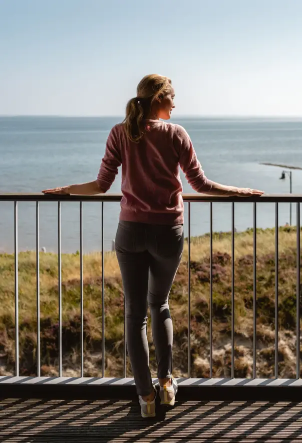 Eine Frau steht auf einem Balkon und blickt auf die Dünen und die Nordsee.