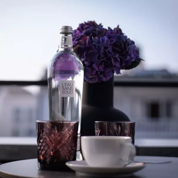 Ein kleiner, runder Tisch auf einem Balkon, auf dem zwei roséfarbene Teelichter, eine dunkle Vase mit lilafarbenem Rhododendron, eine Flasche Wasser und eine weißen Tasse platziert sind.  