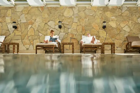Zwei Personen entspannen in weißen Bademänteln auf Liegen am Rand eines Indoor-Pools vor einer Natursteinwand – eine liest, die andere ruht mit geschlossenen Augen.