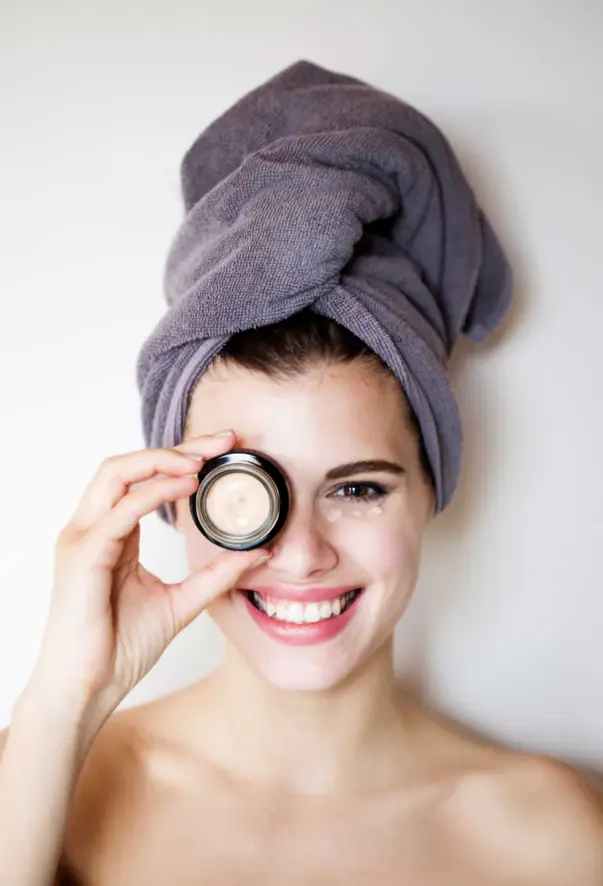 A cheerful woman with a towel around her head smilingly holds a tin of face cream in front of her eye. A few blobs of cream have already been applied under her eye, indicating a skincare routine. The focus is on her radiant smile and the cream, which radiates a positive and caring atmosphere.