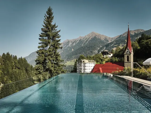 Ein langer Outdoor Infinity-Pool mit einer Berglandschaft mit tannen und einem Kirchturm im Hintergrund.