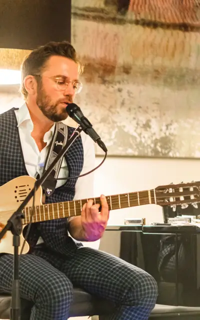 Ein Mann in kartiertem Anzug sitzt auf einem Hocker, spielt auf seiner Gitarre und singt gefühlvoll in eine Mikrofon. 