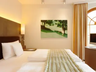 Ein helles Schlafzimmer mit einem Doppelbett, auf dem eine grüne Tagesdecke liegt.  Im Hintergrund hängt ein grünes Bild an der Wand und es ist ein Fenster zu sehen, welches von einer gelben Gardine geschmückt wird.