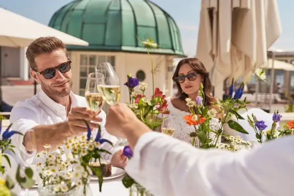 Eine Gruppe von drei Leuten sitzt an einem mit vielen Blumen verzierten und dekorierten Tisch auf einer Terrasse. Die Sonne scheint und im Hintergrund ist die Kulisse von Binz zu sehen. Zwei der Personen stoßen mit ihren Sektgläsern an. 