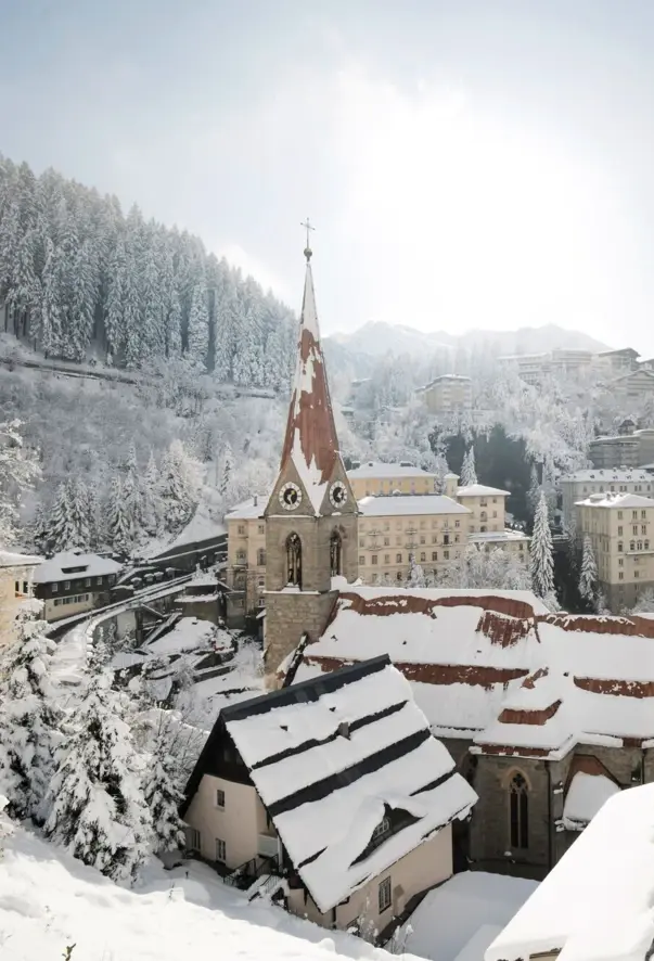 Schneebedeckte Stadt mit Häusern, einer Kirche und einem Berg im Hintergrund an einem klaren Wintertag.