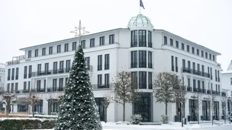 Winterliche Szene mit einem Gebäude und einem verschneiten Baum davor.