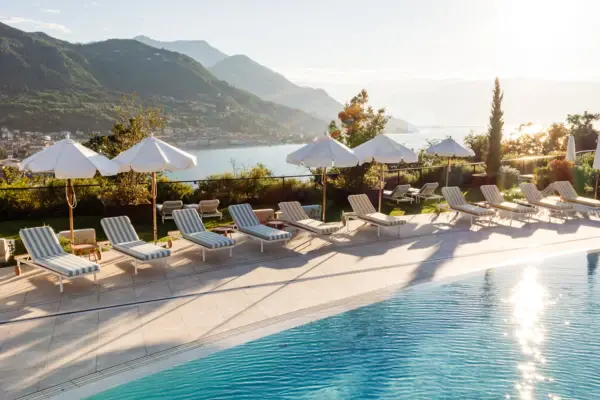 Piscina con sedie e ombrelloni accanto a uno specchio d'acqua, circondata da alberi e montagne.