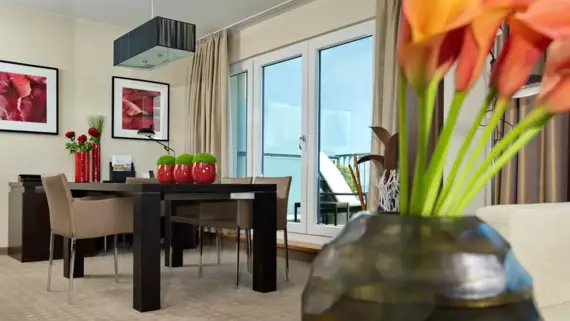 Ein großer dunkler Tisch mit Stühlen steht in einem hellen Hotelzimmer. Es sind verschiedene Pflanzen und Blumen Dekorationen zu sehen sowie ein Durchgang zu einer Terrasse im Hintergrund.