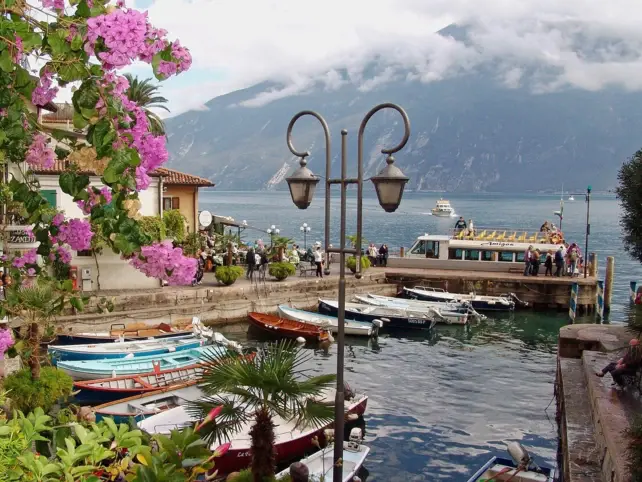 Ein kleiner italienischer Hafen mit Booten, einer Laterne, pinken Blüten und Bergen im Hintergrund.