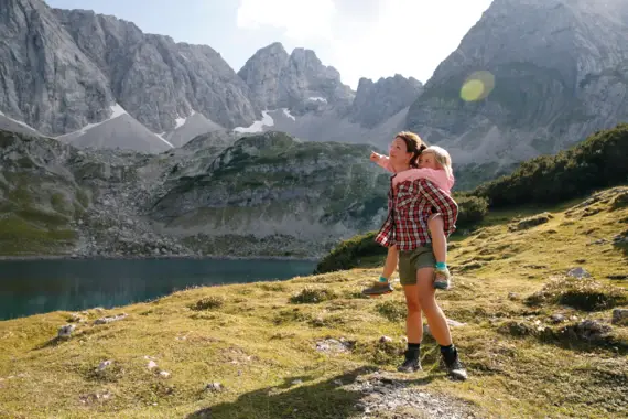 Ein Mann steht mit seiner Tochter auf dem Rücken an einem See in den Bergen. 