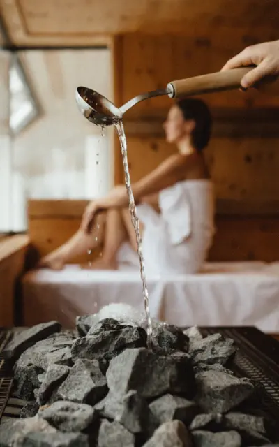 Ein Aufguss wird in einer Sauna gemacht, wobei Wasser aus einer Holzkelle über heiße Steine gegossen wird, was Dampf erzeugt. Im Hintergrund sitzt entspannt eine Person, umhüllt von einem Handtuch, in einer hölzernen Saunalandschaft.
