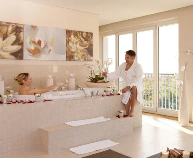 Ein Paar genießt die Ruhe im privaten SPA. Die Frau liegt in der schaumigen Badewanne und der Mann stößt mit ihr mit einem Glas Weißwein an. 