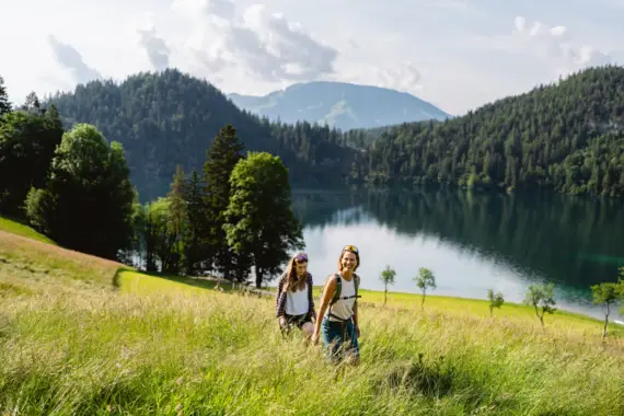 Zwei Frauen wandern durch eine saftig grüne Wiese mit Blick auf einen ruhigen See, umgeben von bewaldeten Hügeln und Bergen im Hintergrund.