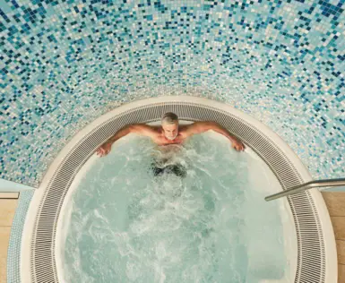 Ein Mann sitzt in einem sprudelnden Whirlpool. Die Wand ist mit kleinen blauen Fliesen dekoriert. 