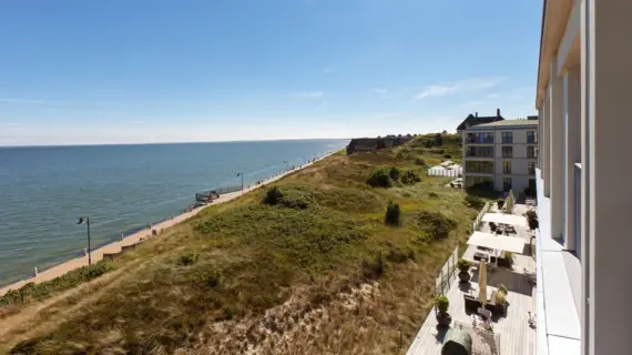 Ein Ausblick auf das Meer, eine Promenade und eine Dünenlandschaft. Vor dem Haus, aus dem das Foto aufgenommen wurde, ist eine Terrasse mit Sitzmöglichkeiten und Sonnenschirmen zu sehen.