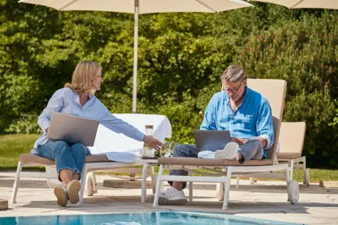 Ein Mann und eine Frau sitzen auf Liegestühlen im Freien und arbeiten an Laptops.