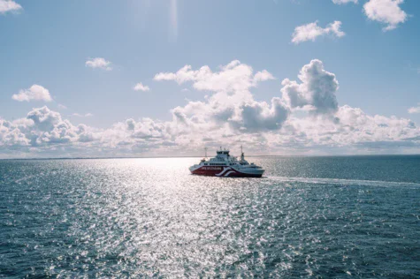 Rot-weißes Fährschiff auf glitzerndem Meer bei Tageslicht mit wolkenbedecktem blauem Himmel im Hintergrund.
