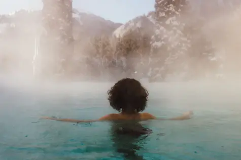 Eine Frau schwimmt in einem warm dampfenden Außenpool mit schneebedeckter Landschaft im Hintergrund. 