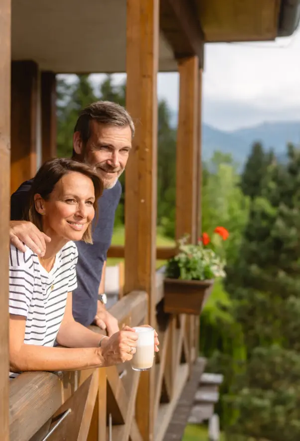 Ein Mann und eine Frau lächeln stehend auf einer Veranda, umgeben von Bäumen.