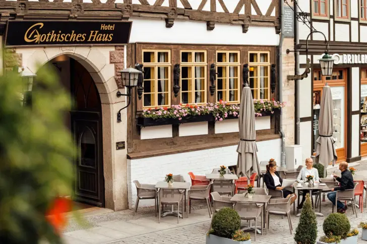 Außenansicht des Hotels Gothisches Haus in Wernigerode mit gemütlicher Terrasse und Fachwerkfassade. Gäste genießen bei Blumen und Sonnenschirmen Kaffee in historischer Altstadtatmosphäre.