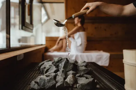 Eine Hand gießt Wasser aus einer Kelle über heiße Steine, während im Hintergrund eine Frau in einem weißen Handtuch auf einer Holzbank sitzt und die wohlige Atmosphäre genießt.