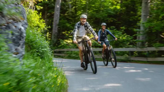 Das Bild zeigt zwei Mountainbiker, die auf einer asphaltierten Straße durch einen dichten Wald fahren. Beide tragen Fahrradhelme und lächeln, während sie Seite an Seite fahren, was auf eine freudige und entspannte Stimmung hindeutet. Die Umgebung ist grün und lebhaft, was vermuten lässt, dass das Bild an einem sonnigen Tag aufgenommen wurde. Die Bewegungsunschärfe verleiht dem Foto ein Gefühl von Geschwindigkeit und Dynamik. Es ist eine Szene, die die Freude am Radfahren und das Erleben der Natur auf zwei Rädern einfängt.
